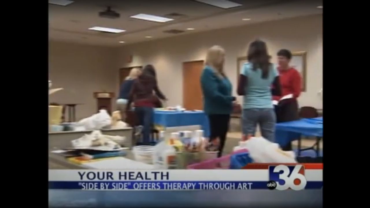 This image shows a still from a television news report. The scene is an indoor setting with several people gathered, some standing and others in motion, possibly interacting or engaged in an activity. There's a table in the foreground with various items spread out, which could be art supplies, indicating the nature of the event related to art therapy as suggested by the news ticker. The news ticker at the bottom of the screen reads "YOUR HEALTH" and "'SIDE BY SIDE' OFFERS THERAPY THROUGH ART" along with the logo of ABC 36. The image appears to be a screenshot from a news broadcast segment.