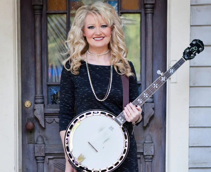 Smiling woman holding a banjo near house entrance.